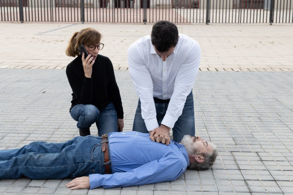 man in cardiac arrest laying on ground while woman calls emergency services and man administers CPR chest compressions
