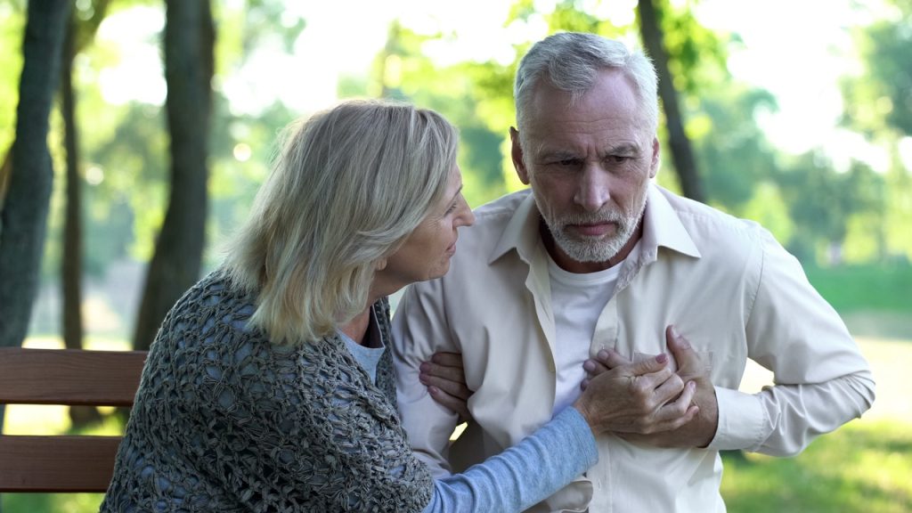 a woman sitting on a bench outdoors concerned about man holding his chest due to pain from heart attack