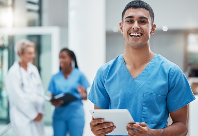 young man in blue scrubs holding tablet with doctor and another nurse in the background
